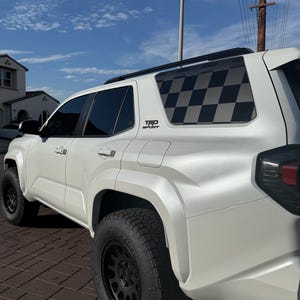 May include: White SUV with black wheels and a checkered flag design on the rear window. The vehicle has the "TRD Sport" emblem and is parked on a brick surface under a blue sky.