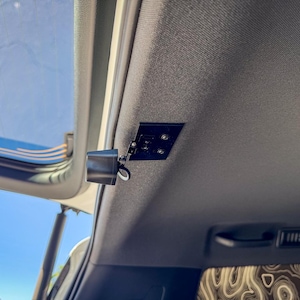 May include: A black camera mounted on the ceiling of a vehicle. The camera is connected to a small black box with wires.