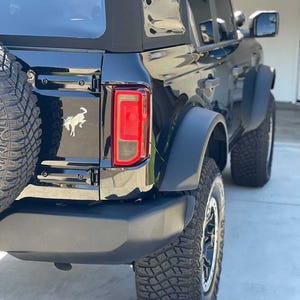 May include: Black SUV with large off-road tires and a spare tire mounted on the back. The vehicle has a black exterior, black fender flares, and a black soft top. The taillight is red. A silver horse emblem is visible on the rear.