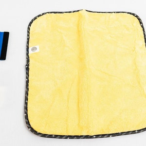 Puede incluir: Un paño de microfibra amarillo con ribete negro y una pequeña etiqueta blanca. El paño está sobre una superficie blanca, junto con una escobilla de goma azul y una botella transparente de líquido.