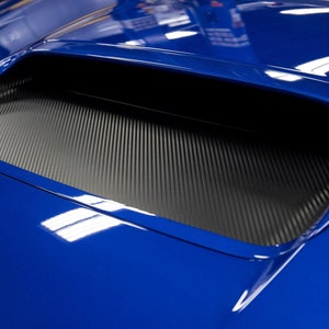 May include: A close-up of a blue car bonnet with a black carbon fibre air intake vent.