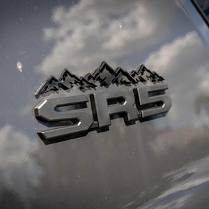 Puede incluir: Primer plano de un emblema negro con las letras SRS y un diseño de montañas. El emblema está montado sobre una superficie gris oscuro, que refleja el cielo y las nubes.