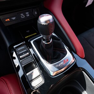 May include: Close-up of a black leather-wrapped gear shifter with a silver knob and red stitching. The shifter is labelled "Type S" in red lettering. The car's interior is black with red accents.