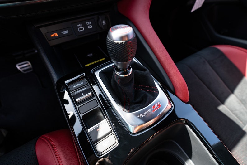 May include: Close-up of a black leather-wrapped gear shifter with a silver knob and red stitching. The shifter is labelled "Type S" in red lettering. The car's interior is black with red accents.