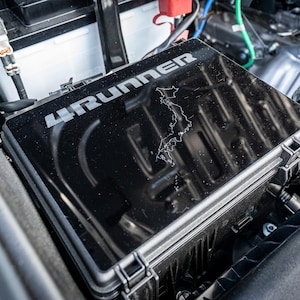 May include: A black plastic box with the words "4RUNNER" printed on the top. The box is open and the inside is empty. The box is sitting in the engine compartment of a car.