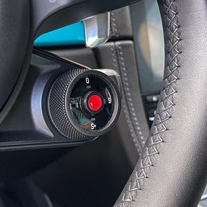 Puede incluir: Primer plano de un volante de cuero negro con costuras blancas. Se puede ver una perilla negra con un botón rojo y los números 0, S y S+.