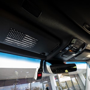 Pode incluir: Um interior de carro preto com um adesivo de bandeira americana branca e cinza na viseira do para-sol. O carro tem um espelho retrovisor e um console de teto preto com botões.