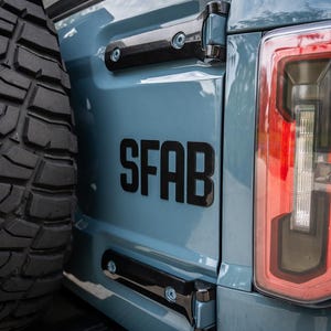 Può includere: Un'auto blu con un pneumatico nero e un adesivo nero "SFAB" sul retro. L'auto ha un fanale posteriore rosso.