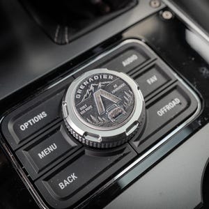 May include: A circular silver-toned dial knob featuring a detailed embossed graphic of a vehicle, mountains, and trees, with the text GRENADIER and BUILT FOR MORE. The knob is positioned on a black console panel with buttons labeled OPTIONS, MENU, BACK, AUDIO, FAV, and OFFROAD.