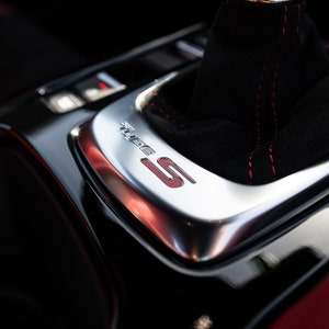 May include: Close-up of a black leather gear shift knob with red stitching and a silver trim plate with the word "Turbo" and the letter "S" in red.