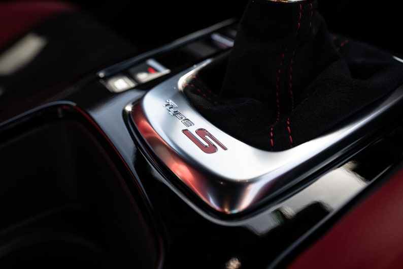 May include: Close-up of a black leather gear shift knob with red stitching and a silver trim plate with the word "Turbo" and the letter "S" in red.