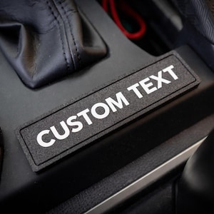 May include: A black rectangular patch with the white text "CUSTOM TEXT" is placed on a black surface, likely inside a vehicle. The patch has a textured appearance, and the surrounding area includes a gear shift and other interior components.