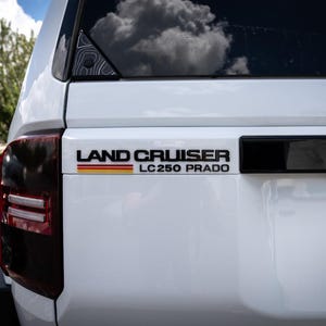 Puede incluir: Land Cruiser LC250 Prado blanco con molduras negras y una franja negra y roja en la parte trasera. El coche tiene un parachoques trasero negro y una matrícula negra con un emblema militar.