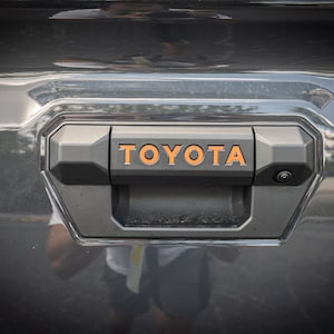 May include: A black tailgate handle with a silver camera lens and an orange "TOYOTA" logo.