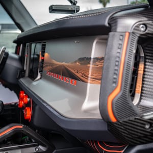May include: A close-up of the interior of a Ford Bronco with a carbon fibre dashboard and a large screen displaying a desert landscape. The screen also displays the word "Bronco" in red lettering. The dashboard has a red and orange accent.
