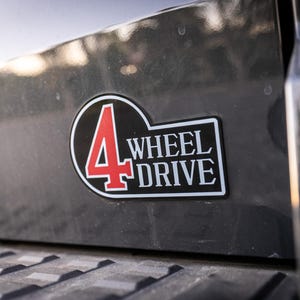 May include: A black and white 4 Wheel Drive sticker with a red "4" on a dark grey surface. The sticker is rectangular with a black border and is affixed to the side of a vehicle.