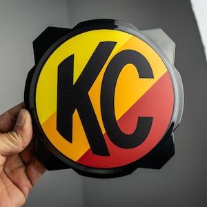Op de afbeelding: Een rond zwart embleem met een geel, oranje en rood streepontwerp. De letters "KC" zijn zwart en gecentreerd op het embleem.