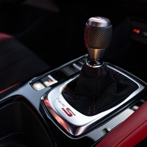 May include: Close-up of a black leather car interior with a silver gear shifter and red stitching. The gear shifter has a black knob with a textured surface and a red "S" logo.