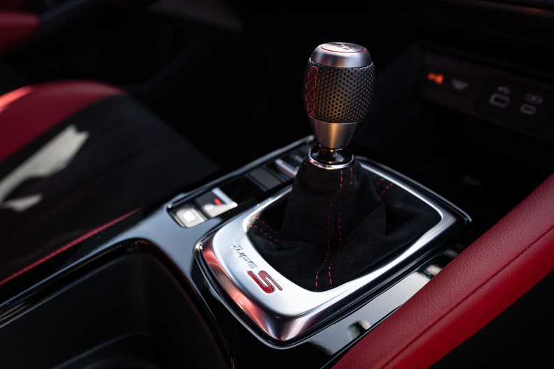 May include: Close-up of a black leather car interior with a silver gear shifter and red stitching. The gear shifter has a black knob with a textured surface and a red "S" logo.