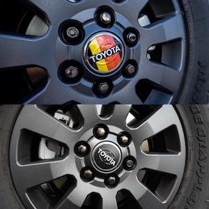 May include: Close-up of two grey car wheels with black lug nuts and a black Toyota emblem. The top wheel has a red, yellow and black emblem with the word "Toyota" on it.