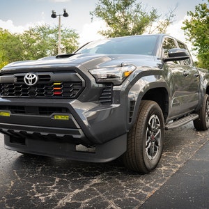 May include: A gray Toyota Tacoma pickup truck with a black grille and yellow accent lights. The truck has a black bumper and black wheels.
