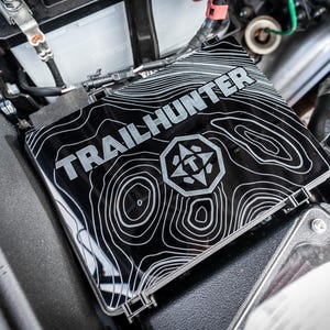 Puede incluir: Cubierta de batería Trailhunter negra y plateada con un diseño de mapa topográfico. La cubierta está instalada en el compartimento del motor de un vehículo, con varios componentes mecánicos visibles en el fondo.