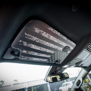 Puede incluir: Un parasol de coche negro con un diseño de bandera americana desgastada en blanco y gris.