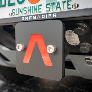 Puede incluir: Una cubierta de enganche cuadrada negra con un logotipo de cheurón rojo en el centro, asegurada por dos pernos hexagonales plateados. El accesorio está montado en la parte trasera de un vehículo bajo una matrícula que dice Sunshine State y Grenadier.