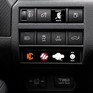 May include: Close-up of a black dashboard with illuminated buttons. The buttons feature various icons and text, including "EJECTO SEAT-O", "AC 120V", "WIFE MUTE", and "KC". The dashboard also has a "TRD LIGHT BAR" label.