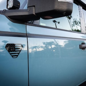 May include: A light blue vehicle door with a black diamond-shaped emblem featuring a black and white American flag design. The door has a black trim and a black door handle. The side mirror and a light are visible.