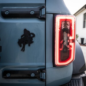 May include: A close-up of the rear taillight of a blue Ford Bronco. The taillight is red and features a graphic of a Bigfoot walking through a forest. The Bronco has a black silhouette of a cowboy riding a horse on the back.