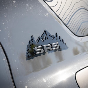 Könnte beinhalten: Nahaufnahme eines grauen Fahrzeugemblems mit dem silbernen Text "SR5", eingebettet in ein Berg- und Kiefern-Design. Das Emblem ist auf einer grauen Oberfläche montiert, mit einem topografischen Kartendesign im Hintergrund.