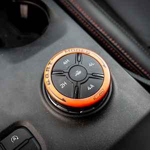 Pode incluir: Close-up de um botão de controle de veículo preto e laranja. O botão é rotulado como "MODOS" e apresenta configurações para 4H, 4L, 2H e 4A, juntamente com o texto "G.O.A.T.". O botão está em um console preto.