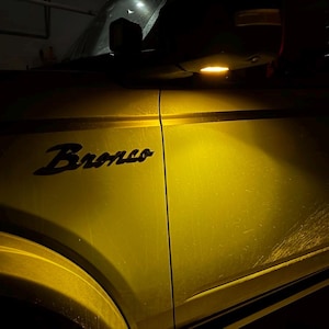 Puede incluir: Un Ford Bronco amarillo con la palabra "Bronco" en letras negras en el lateral. El vehículo tiene un espejo lateral con una luz amarilla.