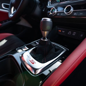 May include: A close-up of a black leather car interior with a red leather trim. The car has a manual transmission with a black leather gear knob and a silver gear selector plate with the letter "S" in red.