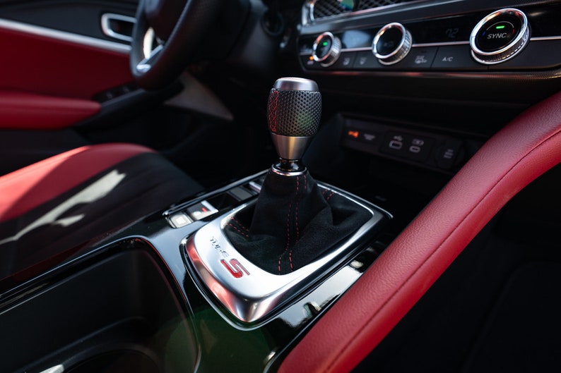 May include: A close-up of a black leather car interior with a red leather trim. The car has a manual transmission with a black leather gear knob and a silver gear selector plate with the letter "S" in red.