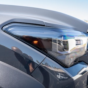 May include: Close-up of a gray car's front headlight with a clear lens and an amber turn signal. The headlight is on and the turn signal is flashing.