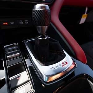 Puede incluir: Primer plano de una palanca de cambios de coche de cuero negro con ribete plateado y costuras rojas. La palanca de cambios tiene una perilla negra con una parte superior plateada y el texto "Type S" en letras rojas.
