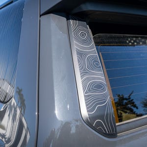 May include: Close-up of a gray vehicle with a window featuring a white topographic map design. The design is repeated on the window frame. The vehicle is parked outside on a sunny day.