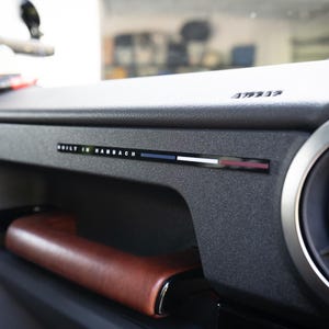 Puede incluir: Un salpicadero de coche negro texturizado con un asa de agarre de cuero marrón y un emblema horizontal con el texto BUILT IN HAMBACH seguido de una franja azul, blanca y roja. Una rejilla de ventilación circular es visible a la derecha.
