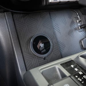 May include: A black Qi wireless charging pad installed in a vehicle's console. The charging pad is round with a silver trim and the Qi logo. The console is black with a geometric pattern.