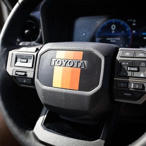 May include: Close-up of a black leather steering wheel with a black and orange stripe and the word "TOYOTA" in silver lettering. The steering wheel has buttons for controlling the radio, cruise control, and other vehicle functions.