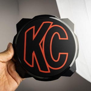 Könnte beinhalten: Rundes schwarzes Emblem mit einem roten "KC"-Logo. Das Emblem hat eine strukturierte, leicht erhabene Oberfläche.