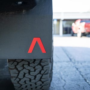 May include: Black mud flap with a red, stylized "A" logo, mounted on a vehicle. The image shows a portion of a black tire with a rugged tread pattern. The background is blurred, suggesting an outdoor setting.