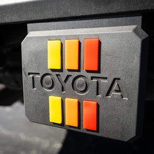 May include: Black rectangular Toyota hitch cover with six vertical stripes in yellow, orange, and red. The word "TOYOTA" is embossed in white letters. The hitch cover is mounted on a vehicle.