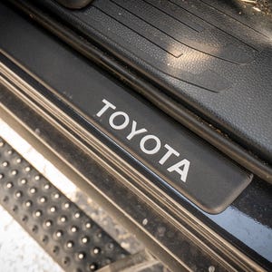 May include: A black Toyota door sill plate with the brand name in white capital letters. The plate is surrounded by black textured plastic and a metal step with small, round, raised grips.