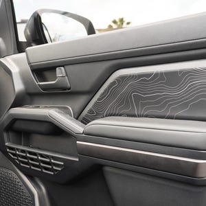 Puede incluir: Panel de puerta de coche gris oscuro con un diseño de mapa topográfico. La puerta incluye un tirador negro, un reposabrazos y un compartimento inferior. El panel tiene un diseño moderno y robusto.