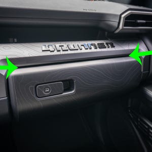 以下が含まれることがあります： シルバーの文字で「4Runner」と書かれた車のダッシュボードの詳細。ダッシュボードは濃いグレーの地形パターンです。プッシュボタンラッチ付きのグローブコンパートメントが見えます。