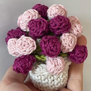 Könnte beinhalten: Ein gehäkelter Blumenstrauß in einem weißen Topf. Die Blumen sind rosa und lila.