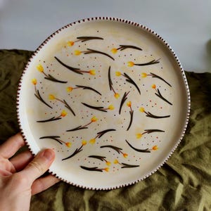 May include: A white ceramic plate with a brown rim and a pattern of yellow flowers and brown stems on a cream background.